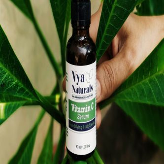 Hand holding Vya Naturals Vitamin C Serum bottle with green leaves background.