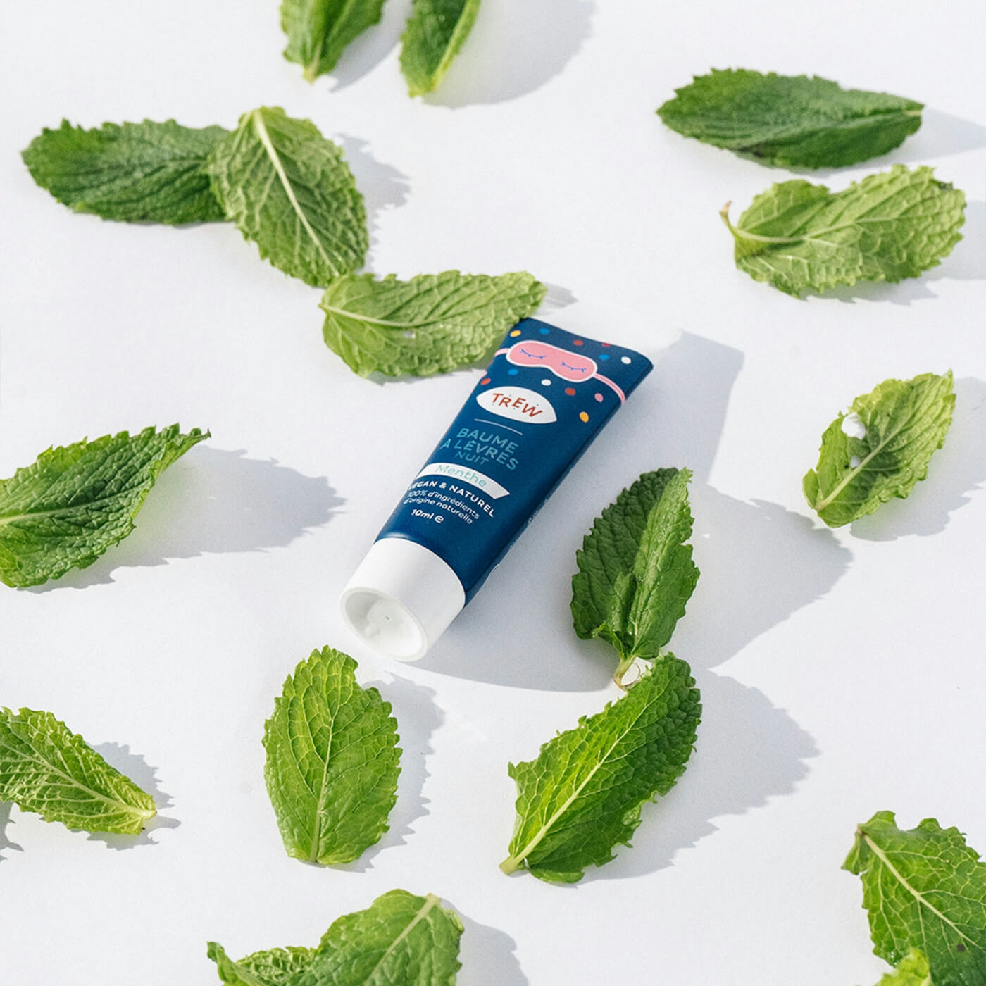 A tube of Trew balm surrounded by fresh green leaves on a white surface.