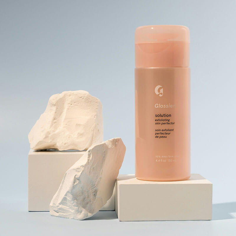 Glossier exfoliating skin perfector bottle displayed with white textured stones.