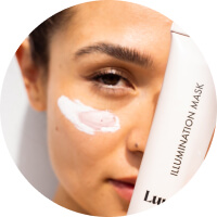 Close-up of a woman with luminous skin holding an illumination mask product.